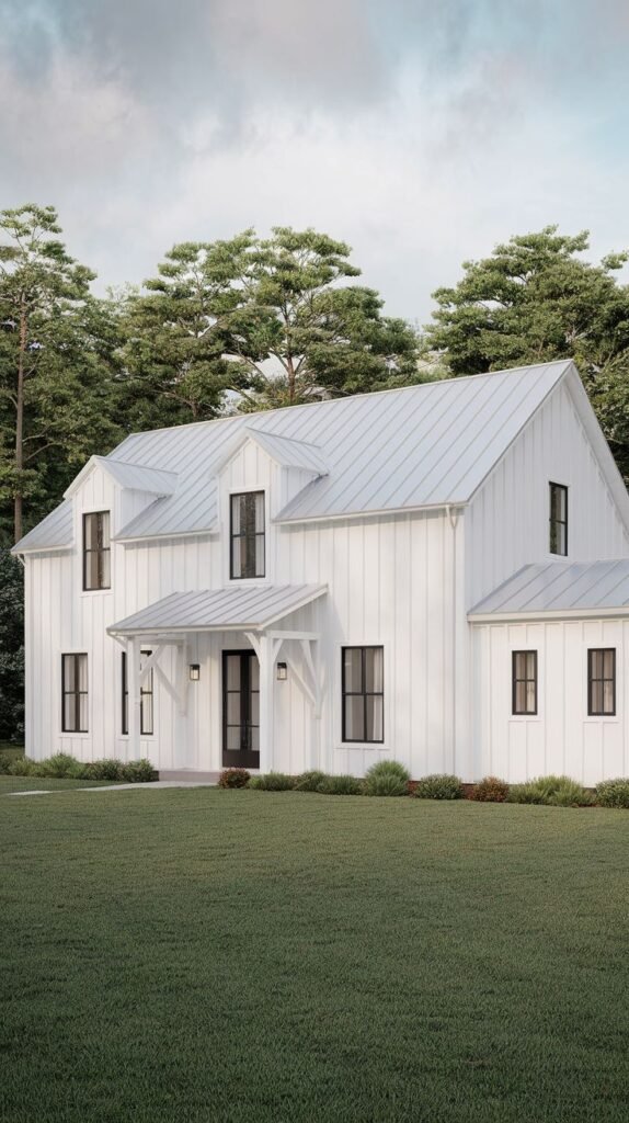 Angled view of a complex white two-story farmhouse structure. The house features multiple gables, a light gray metal roof, and a small projecting entry porch over the black front door, set on a green lawn bordered by trees.