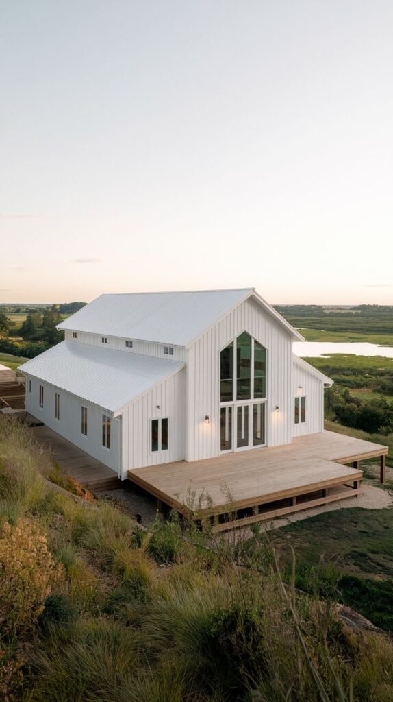 Large white gabled farmhouse positioned on a grassy hill overlooking a distant body of water. The facade features a soaring A-frame window wall and a substantial light wood deck extension.