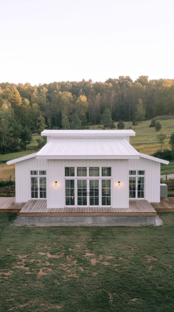Symmetrical single-story white barn-style home featuring multiple white-framed glass doors across the front, opening onto a raised wooden deck. The house is set against a backdrop of green forest.