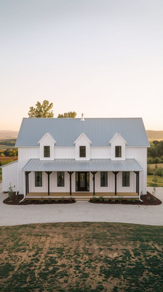 Traditional two-story white farmhouse with a long, covered front porch supported by dark posts. The porch sits beneath a light gray metal roof featuring three symmetrical dormer windows.